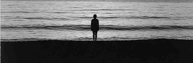 Contact Mallorca Art A grainy, black-and-white, panoramic photo. A person stands silhouetted in the center of the image, facing away from the camera and toward a dark, rippling body of water.